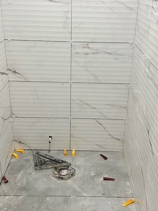 Ribbed white marble-look tile on the walls of a small toilet room, with tile wedges and spacers still on the grey floor